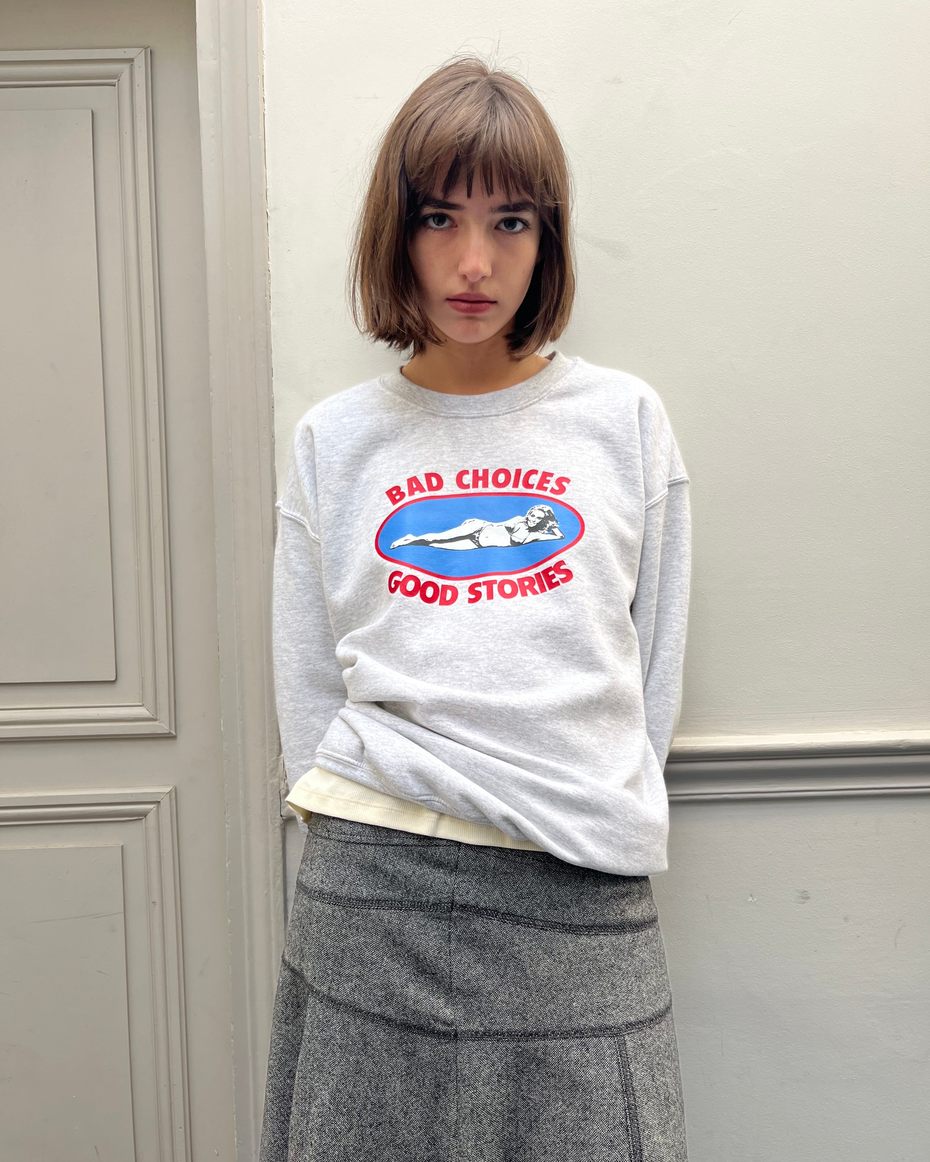 Person wearing a gray sweatshirt with a graphic design and text, standing against a plain wall.