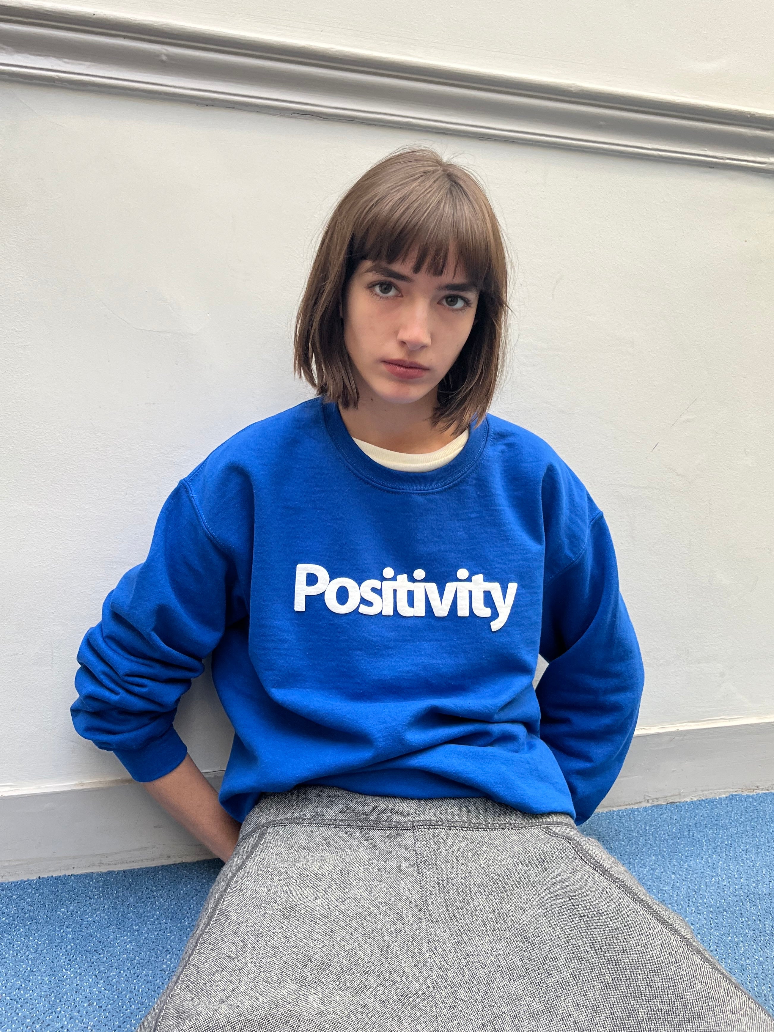 Person wearing a blue sweatshirt with 'Positivity' text against a plain wall.