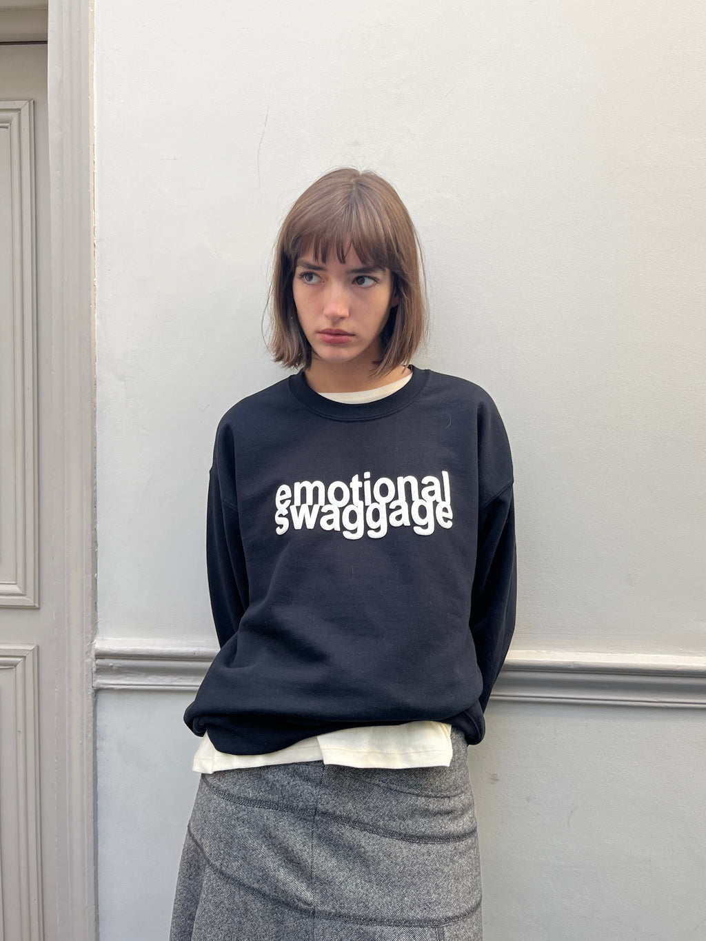 Person wearing a black sweatshirt with 'emotional swaggage' text against a light gray wall.
