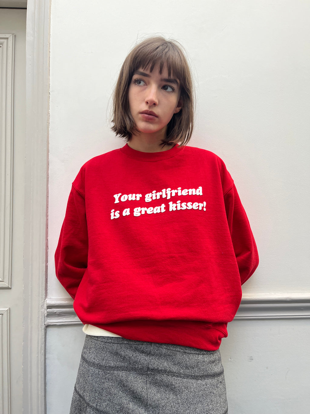 Person wearing a red sweatshirt with text, standing against a white wall.